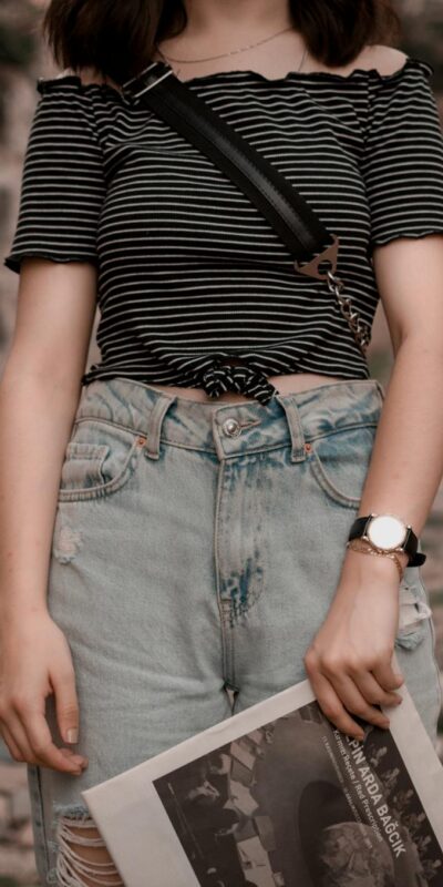 Stylish woman wearing denim jeans and striped top in rustic outdoor setting, holding a magazine.