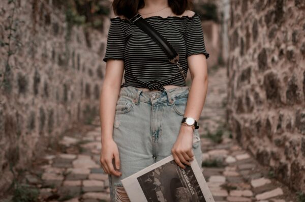 Stylish woman wearing denim jeans and striped top in rustic outdoor setting, holding a magazine.