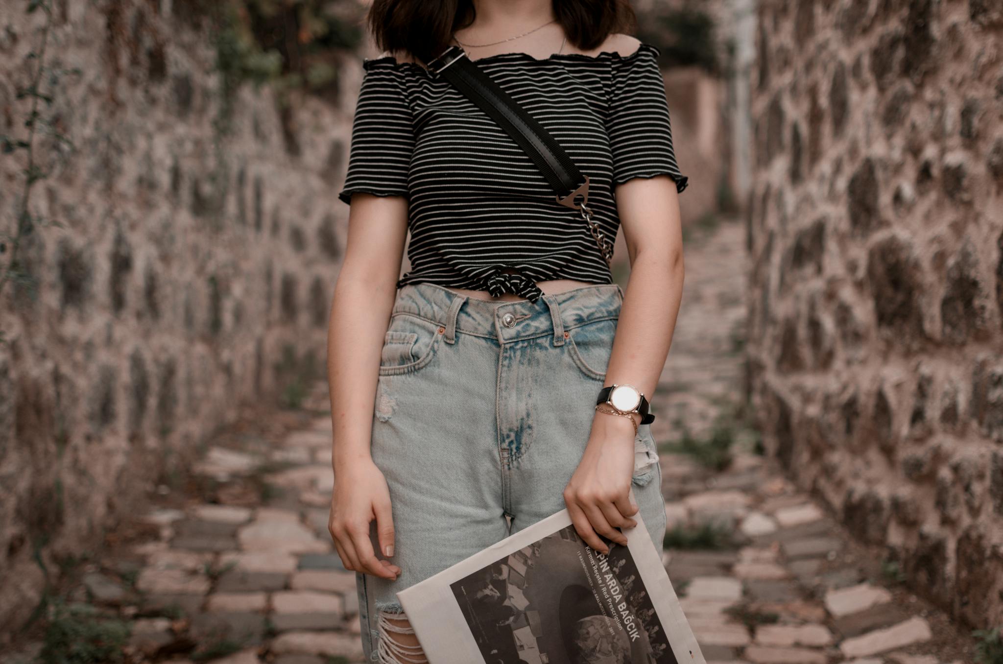 Stylish woman wearing denim jeans and striped top in rustic outdoor setting, holding a magazine.